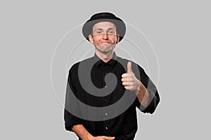 Portrait of a man in a black shirt and pork pie hat holding thumb up isolated over grey background