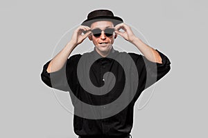 Portrait of a man in a black shirt, pork pie and sun glasses hat isolated over grey background.