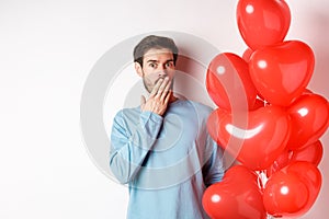 Portrait of man boyfriend standing near Valentines day heart balloons and gasping shocked, standing over white