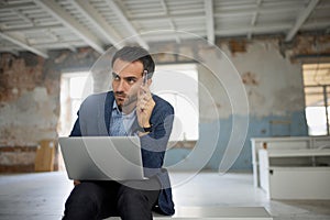 Portrait of man, businessman, worker sitting with laptop with thoughtful expression in big empty room. Thinking process