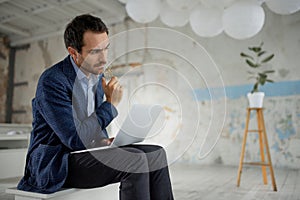 Portrait of man, businessman, worker sitting with laptop with thoughtful expression in big empty room. Working on