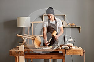 Portrait of man carpenter wearing brown apron and black cap using mallet and chisel in his workshop surrounded by tools and