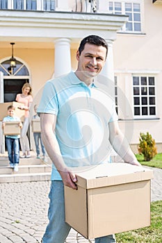 Portrait of man carrying cardboard box while moving house with family in background