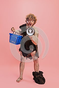 Portrait of man in character of neanderthal, primitive person shouting in megaphone and holding shopping basket isolated