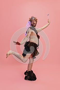 Portrait of man in character of neanderthal wearing fairy wings and crown posing like little wizzard isolated over pink
