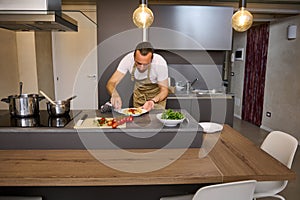 Portrait of a man in chef& x27;s apron, standing at kitchen island and using kitchen towel, wiping away traces of tomato