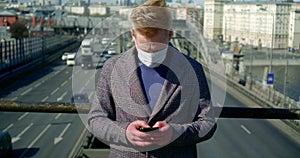 Portrait of a man on a city street. he puts a disposable mask on his face, lowers his head, and points at his smartphone