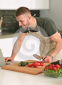 Portrait of man cooking vegetable in the kitchen while looking at a laptop computer on the table