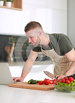 Portrait of man cooking vegetable in the kitchen while looking at a laptop computer on the table