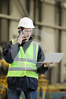 Portrait of a man , factory engineer in work clothes holding laptop and phone, controlling the work process