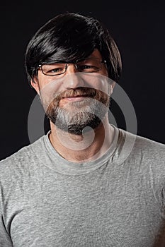 Portrait of a man with grey and black beard and long black hairs. Black background. Male dressed in grey t-shirt, wearing glasses