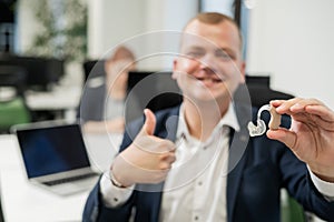 Portrait of a man holding a hearing aid and showing thumbs up in the office.