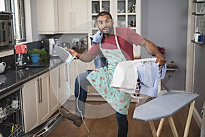 Portrait of man holding laundry basket and iron while closing dishwasher in kitchen