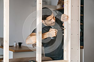 Portrait of a man in home clothes with a screwdriver in his hand fixes a wooden construction for a window in his house. Repair