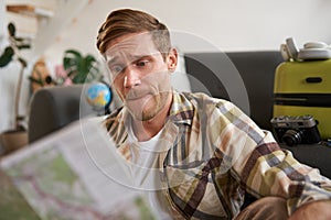 Portrait of man looking confused at travel map, choosing route, going on holiday, preparing for vacation, sitting in