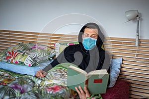 Portrait of man lying and reading a book in bed with sickness and facial mask Covid-19