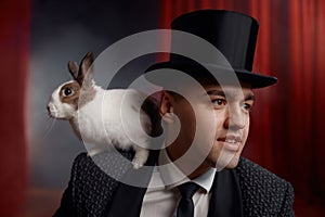 Portrait of man magician with rabbit on shoulder over drapery decorated stage