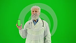 Portrait of man medic on chroma key green screen. Close up senior doctor in white coat holding smartphone with workspace