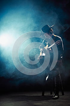 Portrait of man with moustaches in country style clothes playing guitar isolated over dark blue background with smoke