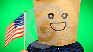 Portrait of man with paper bag on head waving American flag with smiling face against green background.