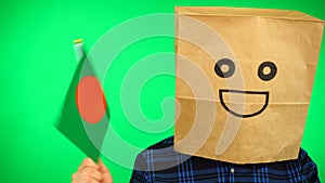 Portrait of man with paper bag on head waving Bangladeshi flag with smiling face against green background.