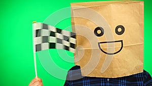 Portrait of man with paper bag on head waving Checkered race flag with smiling face against green background.