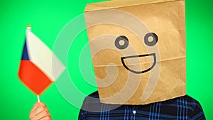 Portrait of man with paper bag on head waving Czech flag with smiling face against green background.