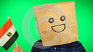 Portrait of man with paper bag on head waving Egyptian flag with smiling face against green background.