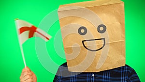 Portrait of man with paper bag on head waving English flag with smiling face against green background.