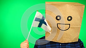 Portrait of man with paper bag on head waving Finnish flag with smiling face against green background.