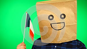 Portrait of man with paper bag on head waving French flag with smiling face against green background.