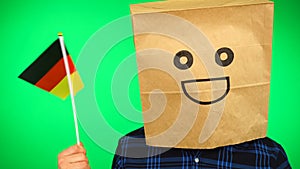 Portrait of man with paper bag on head waving German flag with smiling face against green background.