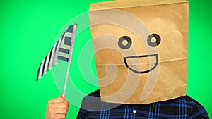 Portrait of man with paper bag on head waving Greek flag with smiling face against green background.