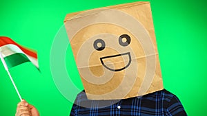 Portrait of man with paper bag on head waving Hungarian flag with smiling face against green background.