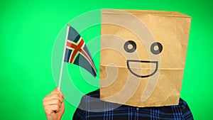 Portrait of man with paper bag on head waving Icelandic flag with smiling face against green background.
