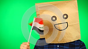 Portrait of man with paper bag on head waving Iraqi flag with smiling face against green background.