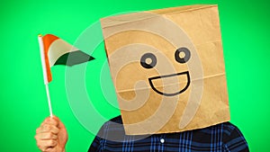 Portrait of man with paper bag on head waving Irish flag with smiling face against green background.