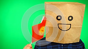 Portrait of man with paper bag on head waving Kyrgyzstan flag with smiling face against green background.