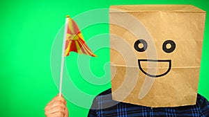 Portrait of man with paper bag on head waving Macedonian flag with smiling face against green background.