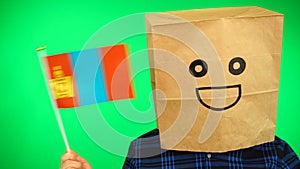 Portrait of man with paper bag on head waving Mongolian flag with smiling face against green background.