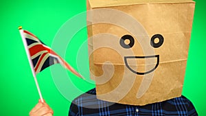 Portrait of man with paper bag on head waving Union Jack flag with smiling face against green background.