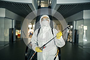 Portrait of a man in a sanitizing disifection suit holding spray near the escalator in an empty shopping mall. A volunteer