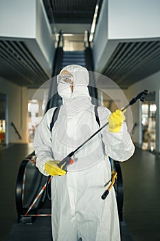 Portrait of a man in a sanitizing disifection suit holding spray near the escalator in an empty shopping mall. A volunteer