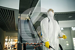 Portrait of a man in a sanitizing disifection suit holding spray near the escalator in an empty shopping mall. A volunteer