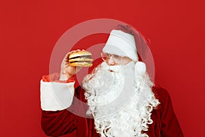 Portrait of man in santa costume standing on red background with burger in hand, hungry looking aside for fast food. Santa will