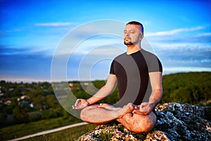 Portrait of the man sitting on a rock in the lotus position against blue sky.