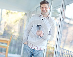 Portrait of man smiling and standing holding a flask. Attractive male leaning against a window inside his home with a