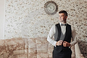 Portrait. A man wearing a white shirt, a black bow tie and black pants and a vest is posing in a room