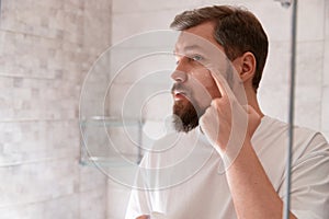 Portrait of man in white tshirt applying cream on his face at bathroom. Self care morning procedure.