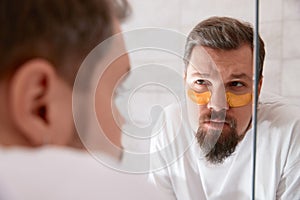 Portrait of man in white tshirt applying eye patches on his face at bathroom. Self care morning procedure